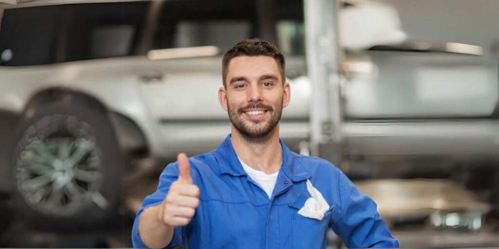 A smiling mechanic in a blue jumpsuit giving a thumbs up inside a garage with a car in the background.