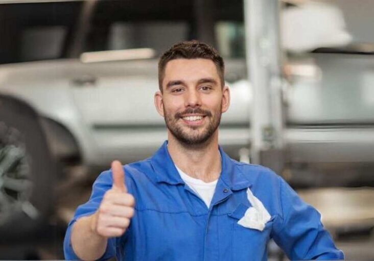 A smiling mechanic in a blue jumpsuit giving a thumbs up inside a garage with a car in the background.